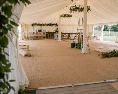 The interior of a marquee mid-setup, showing matting flooring underfoot, a stepladder, and stacked chairs.