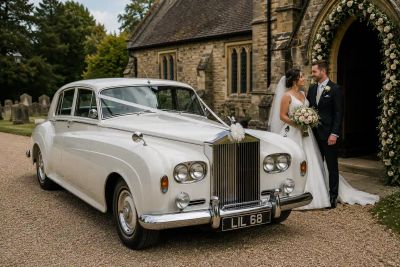 A bride and groom standing beside a vintage white Rolls-Royce wedding car outside a stone church.