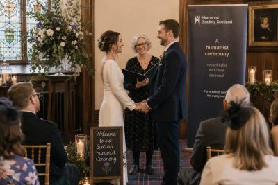 A bride and groom holding hands during a Scottish humanist ceremony, with a Humanist Society Scotland banner and a 'Welcome to our Scottish Humanist Ceremony' sign nearby.