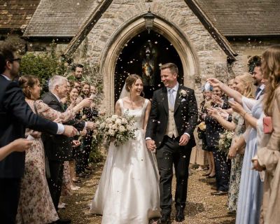 A bride and groom walking hand in hand through confetti thrown by guests outside a stone English church.