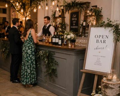 Wedding guests being served at a decorated open bar, with an 'Open Bar, Cheers!' sign listing the evening's drinks.