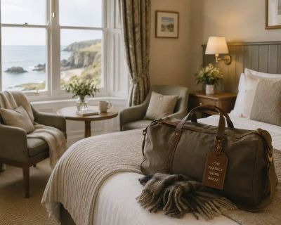 A weekend bag labelled 'The Perfect Short Break' on a hotel bed overlooking a UK coastal view, styled for a minimoon.